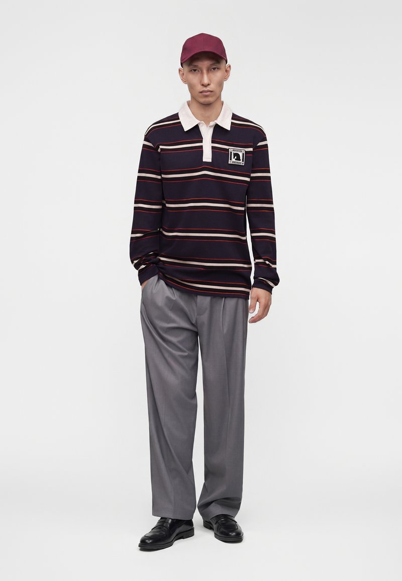 Homme portant une casquette bordeaux, un polo à manches longues noir rayé avec écusson, un pantalon gris à larges jambes et des chaussures habillées noires, debout avec une main dans la poche.