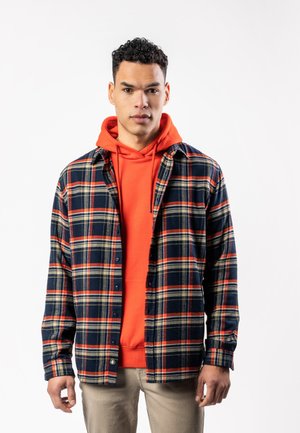 Young man wearing orange hoodie under unbuttoned navy and orange plaid shirt, beige pants, standing against white background.