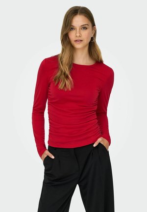 Maglietta rossa a maniche lunghe con dettagli arricciati e scollatura rotonda, abbinata a pantaloni neri a vita alta con pieghe e tasche laterali.