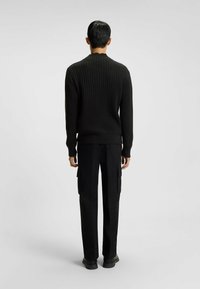 Uomo in piedi, di spalle rispetto all'osservatore, che indossa un maglione a coste nero, pantaloni cargo neri e scarpe nere, su uno sfondo chiaro e uniforme.