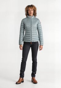 Lichtblauwe gewatteerde jas met een hoge kraag, verticale stiksels en ritssluiting, gecombineerd met zwarte jeans en bruine enkellaarzen.