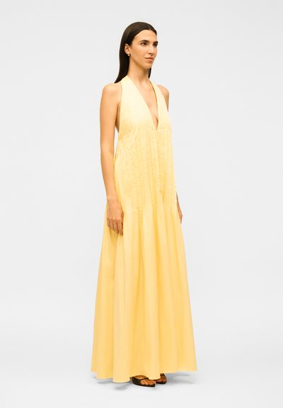 Γυναίκα με μακριά σκούρα μαλλιά, φοράει ένα κίτρινο μάξι φόρεμα χωρίς μανίκια με βαθύ V-λαιμόκοψη και μαύρα σανδάλια, στέκεται μπροστά σε απλό φόντο.