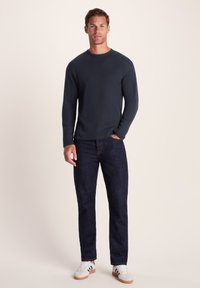 Man standing with hand in pocket wearing dark long-sleeve shirt, dark jeans, and white sneakers against a plain background.