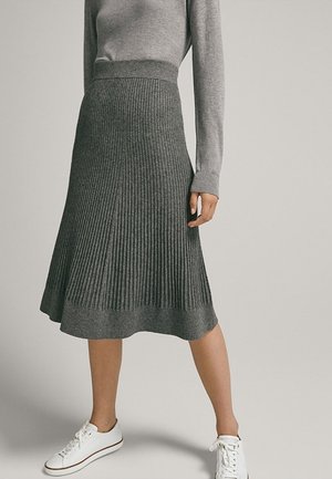 Femme portant une jupe midi en tricot côtelé gris, un haut gris à manches longues et des baskets blanches, debout devant un fond uni.
