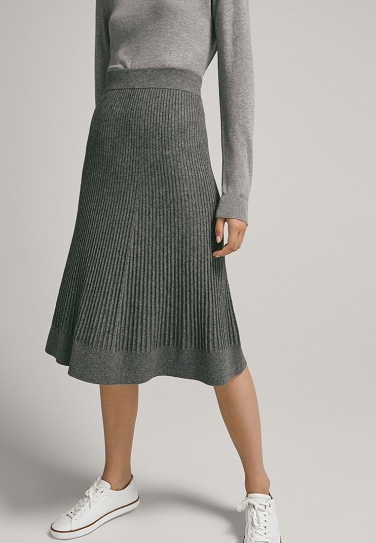Femme portant une jupe midi en tricot côtelé gris, un haut gris à manches longues et des baskets blanches, debout devant un fond uni.