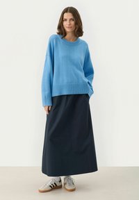 Helles blaues, oversized Sweatshirt mit geripptem Saum, kombiniert mit einem langen schwarzen Rock. Das Modell trägt graue und schwarze Sneakers mit weißen Socken.