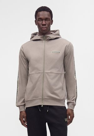 Jeune homme portant un sweat à capuche taupe zippé avec un ruban logo noir sur les manches et un pantalon noir avec un petit logo près de la poche, debout face à l’avant.