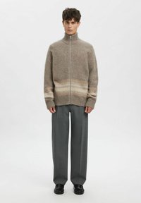 Beige genser med glidelås, strukturert med høy krage og brune gradientdetaljer, kombinert med løstsittende grå bukser og svarte sko.