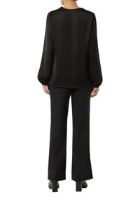 Schwarze Seidenbluse mit langen, geschwollenen Ärmeln, rundem Halsausschnitt und glatter Textur. Kombiniert mit maßgeschneiderten schwarzen Hosen und klobigen Absatzschuhen.