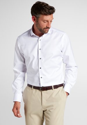 Witte blouse met een verspreide kraag, zwarte knopen en lange mouwen, gedragen met beige broeken en een bruine riem. Soepele textuur.