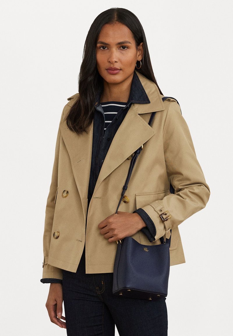 Trench coat bege de duas fileiras de botões dourados, combinado com uma mala cruzada azul-marinho e jeans escuros. Camiseta às riscas visível por baixo.