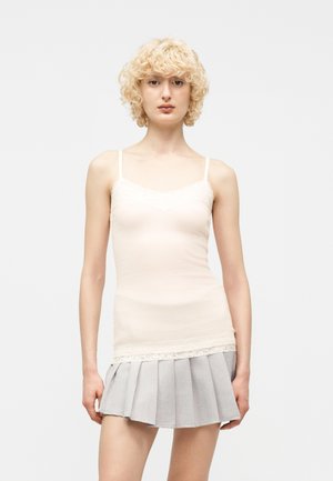 Personne aux cheveux blonds bouclés portant un caraco blanc orné de dentelle et une mini-jupe plissée gris clair, debout devant un fond uni.