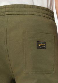 Olíva zöld rövidnadrág, elasztikus derékkal, hátsó zsebbel és egy fekete címkével, amelyen sárga szöveggel az áll: "Workwear". Texturált anyag.