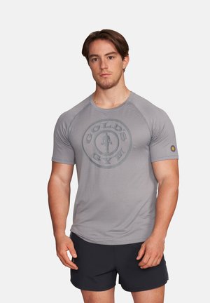 Junger Mann steht und trägt ein enges graues Gold's Gym T-Shirt und schwarze Shorts vor einem einfarbigen hellen Hintergrund.
