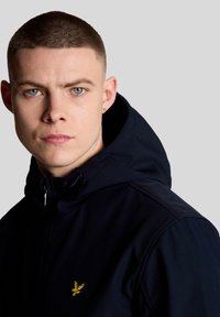 Μπλε σκούρο μπουφάν με κουκούλα, με κίτρινο λογότυπο αετού στο αριστερό μέρος του στήθους. Λείο ύφασμα με κλείσιμο φερμουάρ και ρυθμιζόμενη κουκούλα με κορδόνι.