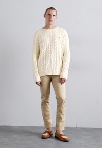 Cream cable-knit sweater, fitted at the waist, paired with light tan chinos and brown slip-on shoes. Smooth texture and ribbed details visible.