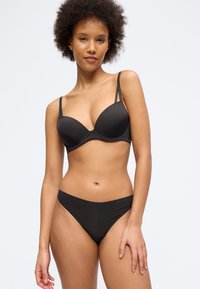 Γυναίκα με σγουρά μαλλιά που φοράει μαύρο σουτιέν με μπανέλα και μαύρο εσώρουχο σε στιλ μπικίνι, στέκεται μπροστά σε απλό φόντο.