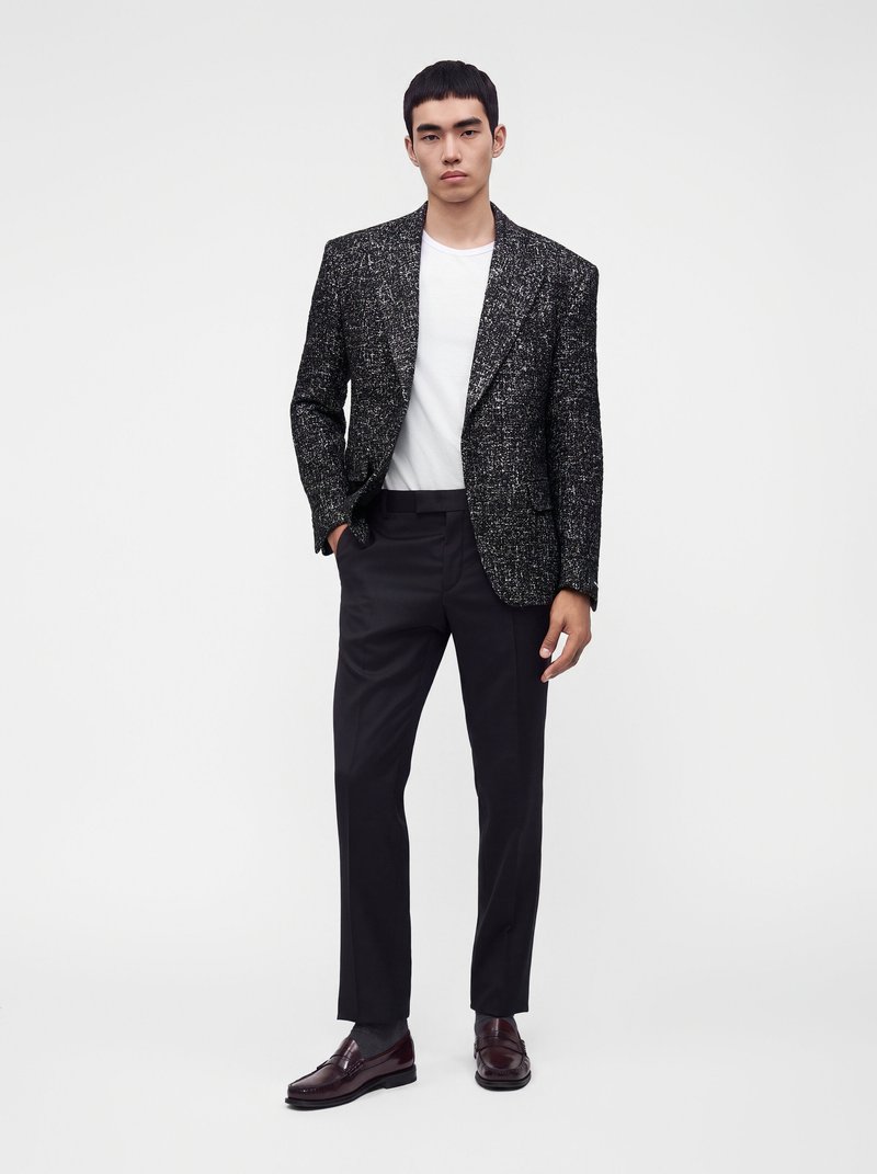 Jeune homme portant un blazer texturé noir et blanc, un t-shirt blanc, un pantalon noir et des mocassins brun foncé, debout devant un fond blanc uni.