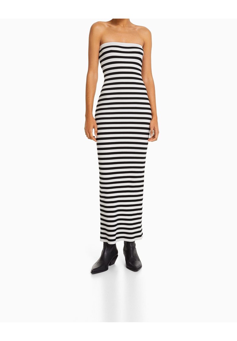 Robe maxi rayée noir et blanc sans bretelles, au design ajusté, avec une texture lisse. Portée avec des bottines noires à la cheville.