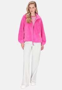 Flauschige, leuchtend pinke Zip-up-Jacke mit Kragen und elastischen Bündchen, kombiniert mit weißen, weit geschnittenen Hosen und einem darunterliegenden gestreiften Oberteil.