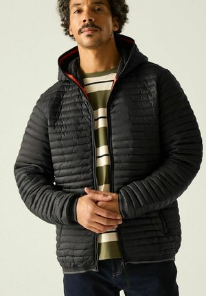 Mann mit Schnurrbart und lockigem Haar, der eine schwarze gesteppte Jacke über einem grün-weiß gestreiften Hemd trägt und mit verschränkten Händen dasteht.