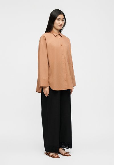 Γυναίκα που φοράει ένα φαρδύ ροδακινί πουκάμισο με κουμπιά, μαύρο παντελόνι με φαρδιά πόδια και δαντελένια τελειώματα, καθώς και καφέ σανδάλια με επίπεδη σόλα, στέκεται μπροστά σε ένα απλό ανοιχτόχρωμο φόντο.