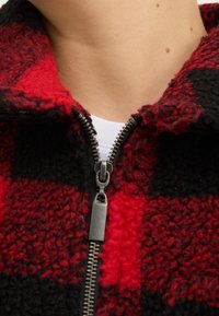 Rød og sort ternet fleecejakke med høj krave, der har en sølvlynlås og blødt, struktureret stof. Nærbillede af halsudskæringen.