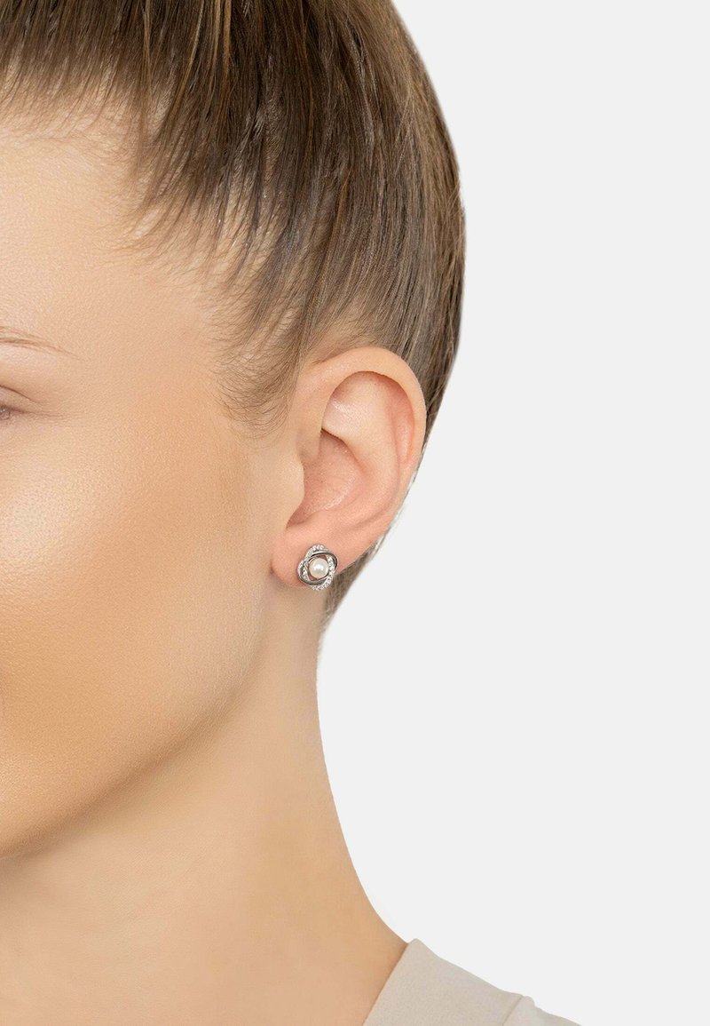 Woman’s ear wearing a silver stud earring with a central pearl surrounded by small crystals against a plain background.