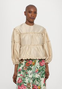 Beige selyemtop ráncokkal, hosszú, bő ujjakkal és magas nyakkal, virágmintás szoknyával párosítva, élénk színekben.