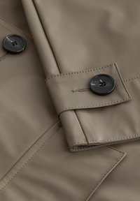 Manche et devant d’un manteau en cuir marron avec des boutons noirs et des coutures détaillées en gros plan.
