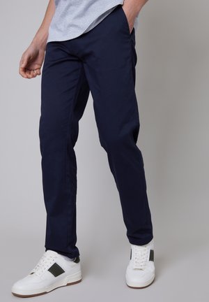 Marineblaue Chinos im Slim-Fit, aus glattem Baumwollstoff, mit Seitentaschen. Kombiniert mit weißen Sneakers mit grünen Akzenten.