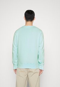 Sudadera verde menta clara con cuello redondo, puños y dobladillo de canalé, hecha de una tela suave. Se lleva con pantalones beige, vista desde la parte posterior.