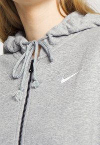 Haut à capuche zippé gris avec un cordon de serrage, présentant un petit logo Nike blanc et un tissu texturé. Les accents incluent des pompons sur les cordons.