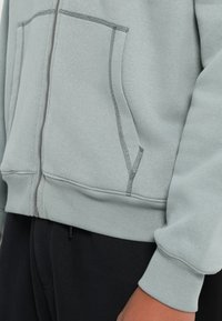 Light gray zip-up sweatshirt with front pockets and ribbed cuffs worn over black pants, shown on a person’s side torso and arm.