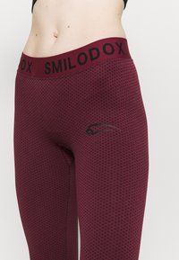 Leggings bordeaux con un motivo a rete testurizzato, una vita nera con la scritta "SMILODOX" e un dettaglio logo discreto di lato.
