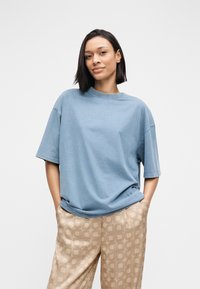 Laisvo silueto mėlyna medvilninė marškinėliai su apvalia iškirpte, trumpomis rankovėmis ir lygiu paviršiumi, derinami su smėlio spalvos raštuotomis kelnėmis.