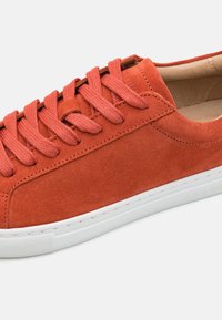 Orange suede sneaker with flat laces, white rubber sole, and a smooth texture. Features minimal stitching and a reinforced toe cap.