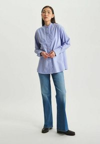 Chemise boutonnée lilas clair avec col montant, manches longues et coupe décontractée, assortie à un jean en denim bleu évasé et des chaussures noires.