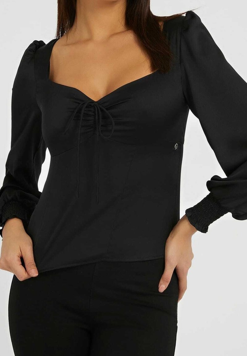 Blouse noire à manches bouffantes, décolleté en forme de cœur, détail froncé à l'avant et poignets élastiqués. Texture lisse avec un léger éclat.
