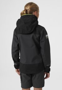 Černá voděodolná bunda s kapucí, s hladkým povrchem a šedými akcenty na manžetách a na ramenou, nošená přes tmavé šortky.