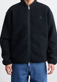 Czarna kurtka fleece z zamkiem z przodu, z wysokim kołnierzem i bocznymi kieszeniami. Haftowane logo na piersi z teksturowanym wykończeniem.