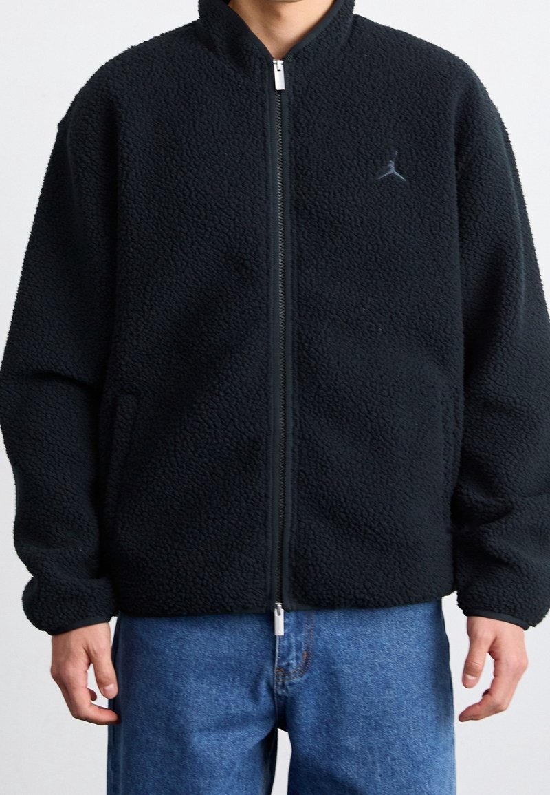 Czarna kurtka fleece z zamkiem z przodu, z wysokim kołnierzem i bocznymi kieszeniami. Haftowane logo na piersi z teksturowanym wykończeniem.