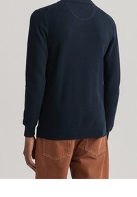 Marineblauer strukturierter Strickpullover mit Kragen, enganliegenden Bündchen und Taillenbund, kombiniert mit braunen Hosen mit kontrastierenden Gesäßtaschen.