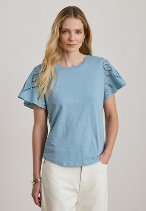 Femme aux longs cheveux blonds portant un haut bleu clair avec des manches volantées en dentelle et un pantalon blanc, debout avec une main dans la poche.