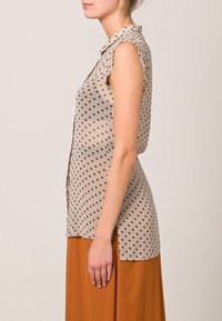 Blusa sin mangas de color beige claro con lunares negros, fabricada en tejido suave, con cuello tipo camisa y un corte relajado, combinada con una falda naranja.