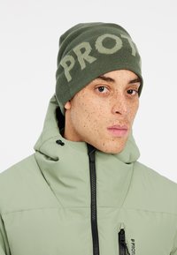 Olivgrüne Strickmütze mit "PRO"-Schriftzug, getragen von einer Person in einer olivgrünen Daunenjacke mit hohem Kragen und Reißverschlusstaschen.