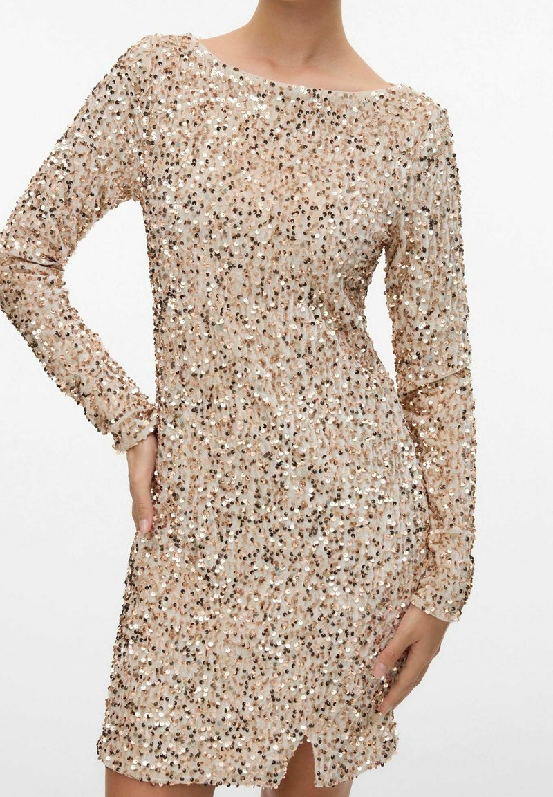 Robe beige à manches longues couverte de sequins brillants, style ajusté avec une petite fente à l'avant, portée par une personne avec une main sur la hanche.