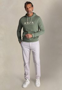 Groene hoodie met crèmekleurige tekst "N.Z.A" en "New Zealand Auckland", gecombineerd met witte broek en witte sneakers. Eenvoudig ontwerp, casual pasvorm.