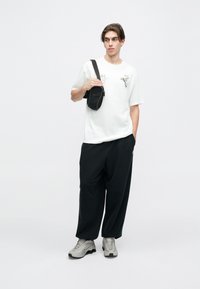 Biely tričko s grafickým dizajnom, čierne voľné nohavice a šedé tenisky. Čierna kabelka cez telo so zipsom je nosená cez hruď.