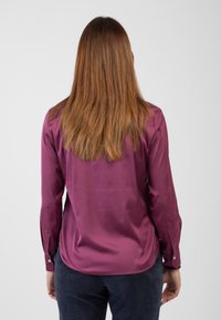 Camicia a maniche lunghe di un profondo colore bordeaux realizzata in tessuto liscio, con polsini abbottonati e orlo arrotondato. Indossata con pantaloni di velluto a coste scuri.